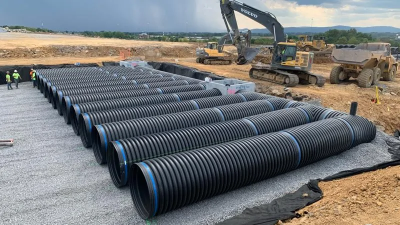 Rows of black corrugated HDPE pipe laid in a gravel bed at a construction site with a Volvo excavator