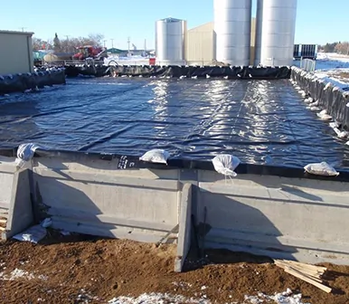 Black geomembrane liner installed in a concrete containment structure with storage silos in the background at a winter jobsite