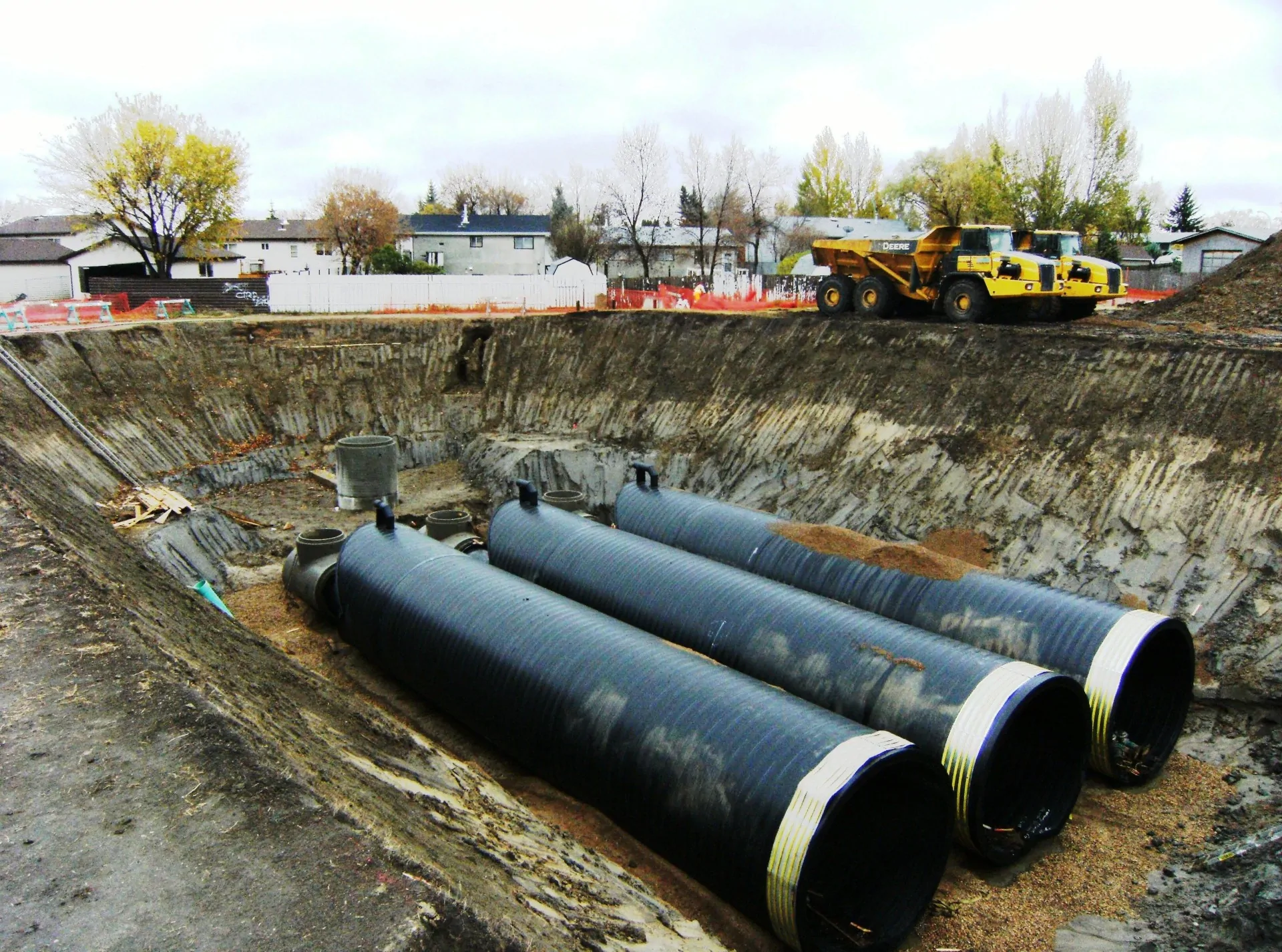 Large-diameter black HDPE pipes staged in an excavated construction pit with a dump truck and houses in the background