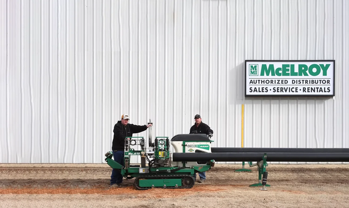 Two workers with a McElroy fusion machine outside a building beside a McElroy authorized distributor sign and black HDPE pipe