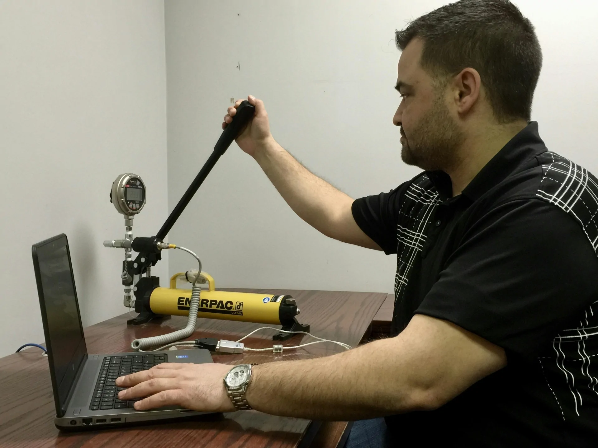 Technician pressure testing with an Enerpac hand pump, a pressure gauge and a laptop at a desk